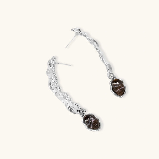 Silver-toned twisted metal earrings with dark brown oval resin stones dangling from sculptural posts on white background.