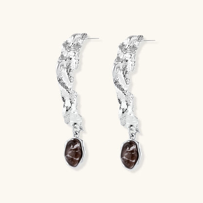 Silver-toned twisted metal earrings with dark brown oval resin stones dangling from sculptural posts on white background.