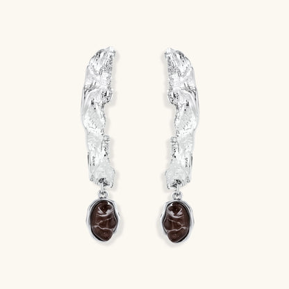 Silver-toned twisted metal earrings with dark brown oval resin stones dangling from sculptural posts on white background.